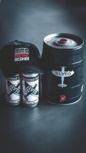 Elevated Seltzer keg that is grey with an airplane logo with the letters ELVTD in it, next to two cans of Elevated Seltzer with a Marcus "Maniac" McGhee hat on top of the hats.